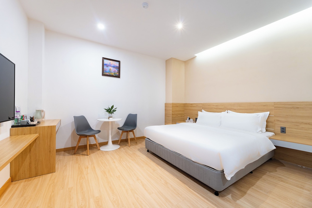 A hotel room with a bed, a desk, two chairs, a small table, and a wall-mounted painting.