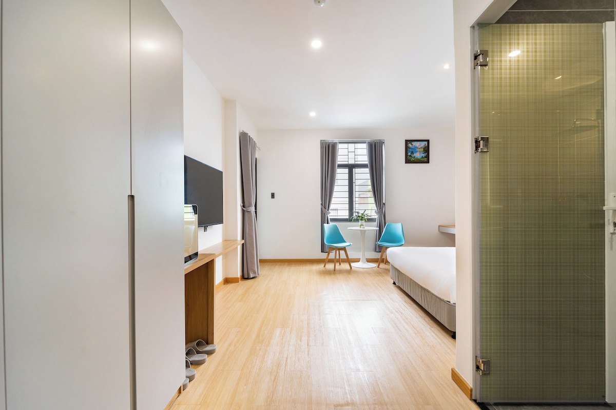 A hotel room with a bed, a desk with a chair, a small table with two chairs, a window with curtains, and a shower area.