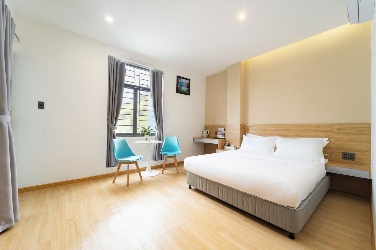 A hotel room with a bed, two chairs, a small table, a window with curtains, and a painting on the wall.