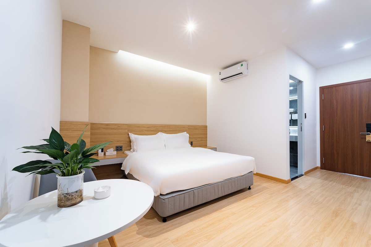 A modern hotel room with a bed, bedside table, potted plant, and a small round table.