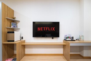 A modern living room with a flat-screen TV mounted on the wall displaying the Netflix logo. There are wooden shelves with decorative items and a small table with a kettle and drinks.