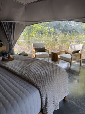 A tented accommodation with a bed, two chairs, a small table, and a view of greenery.