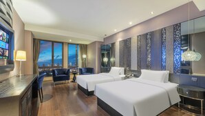 Ein Hotelzimmer mit zwei Betten, einem Schreibtisch, einem Sessel, einem Fernseher und Blick auf die Stadtlandschaft.