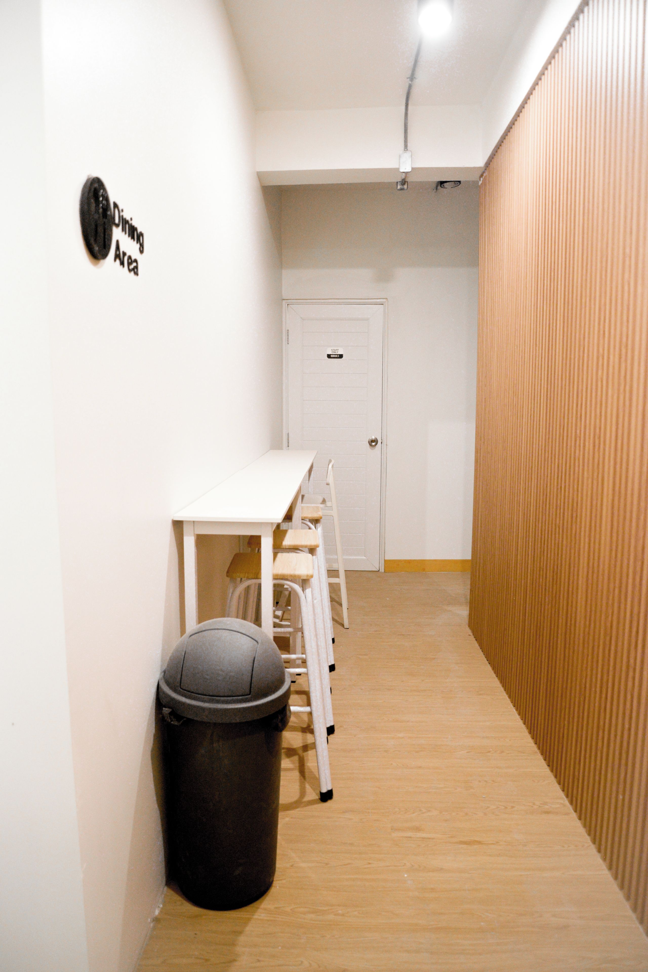 Un couloir étroit avec une porte blanche, une poubelle et une table blanche avec des chaises.