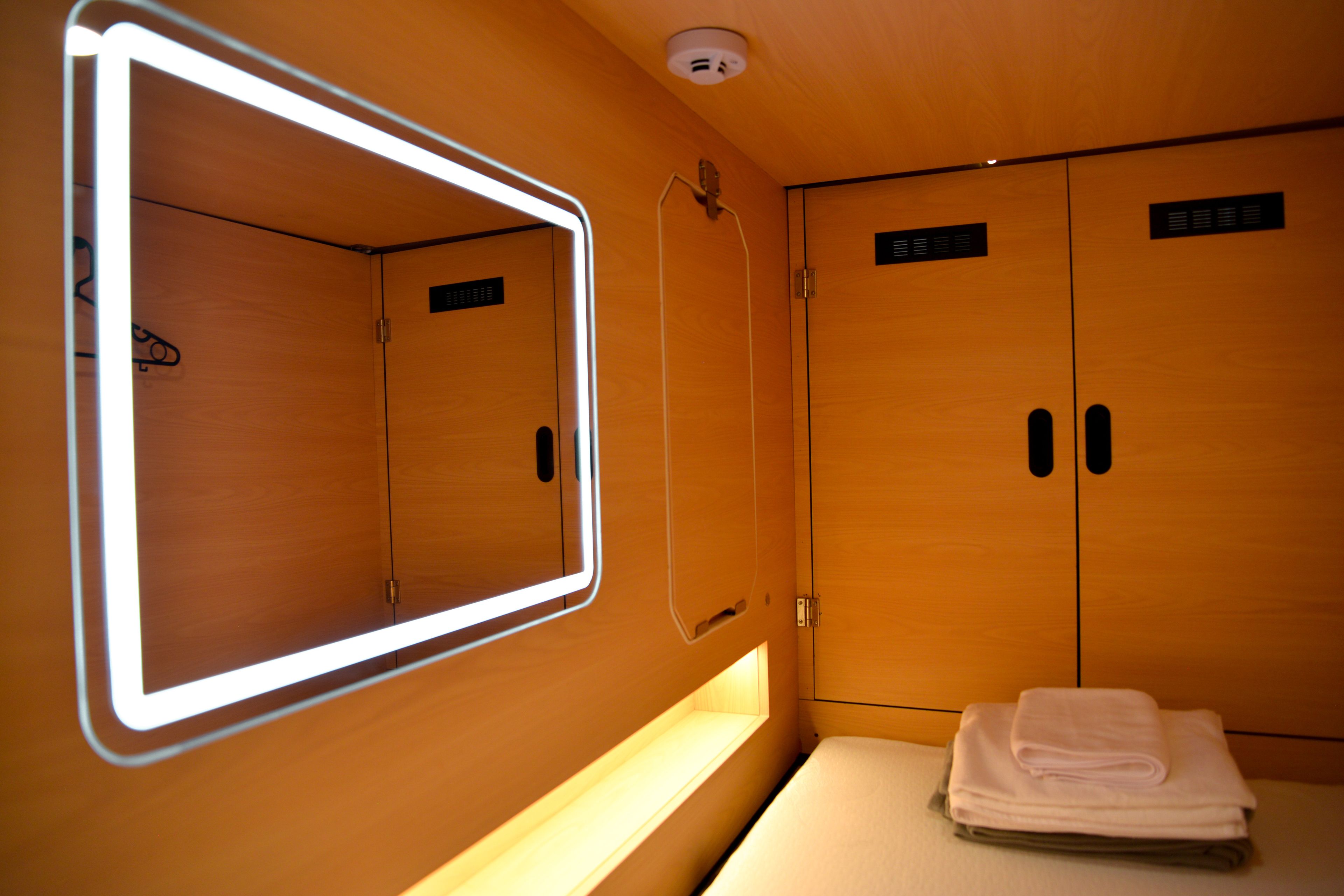 Habitación de hotel con un espejo grande, una tocador con luz, una cama con una toalla blanca doblada con cuidado, y armarios de madera con un letrero pequeño.