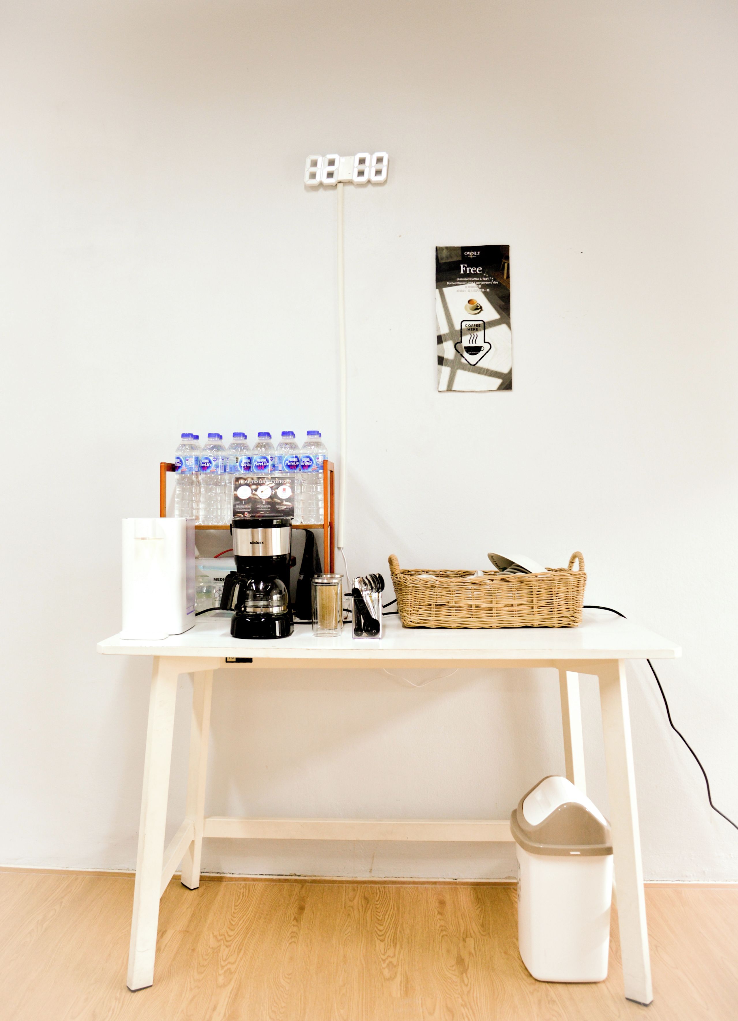 Una mesa blanca con una cafetera, botellas de agua, una cesta y un cubo de basura.