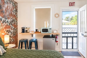 A compact kitchenette with a microwave, coffee maker, and small refrigerator. There are two stools and a bed with a green quilt. An exit sign is visible on the door. - Charming Stay Near Historic District St. Augustine (St. Augustine)