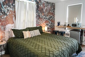 A bedroom with a bed, pillows, a nightstand, a lamp, a wall with a floral mural, a window with curtains, and a kitchenette area with a microwave and refrigerator. - Charming Stay Near Historic District St. Augustine (St. Augustine)