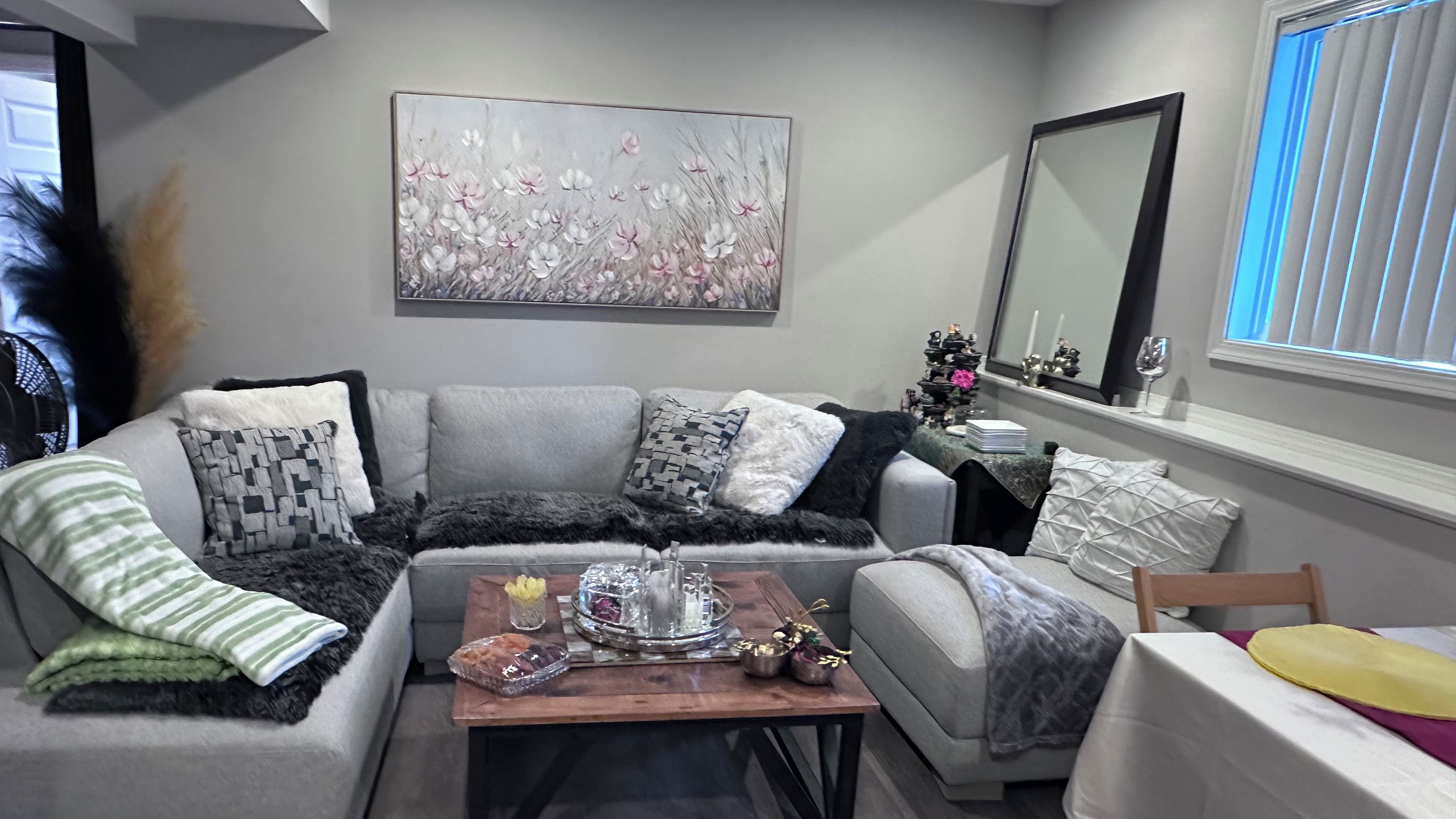 Een moderne woonkamer met een grijze bank, een bijzettafeltje met decoratieve items, een schilderij aan de muur en een raam met zonwering.