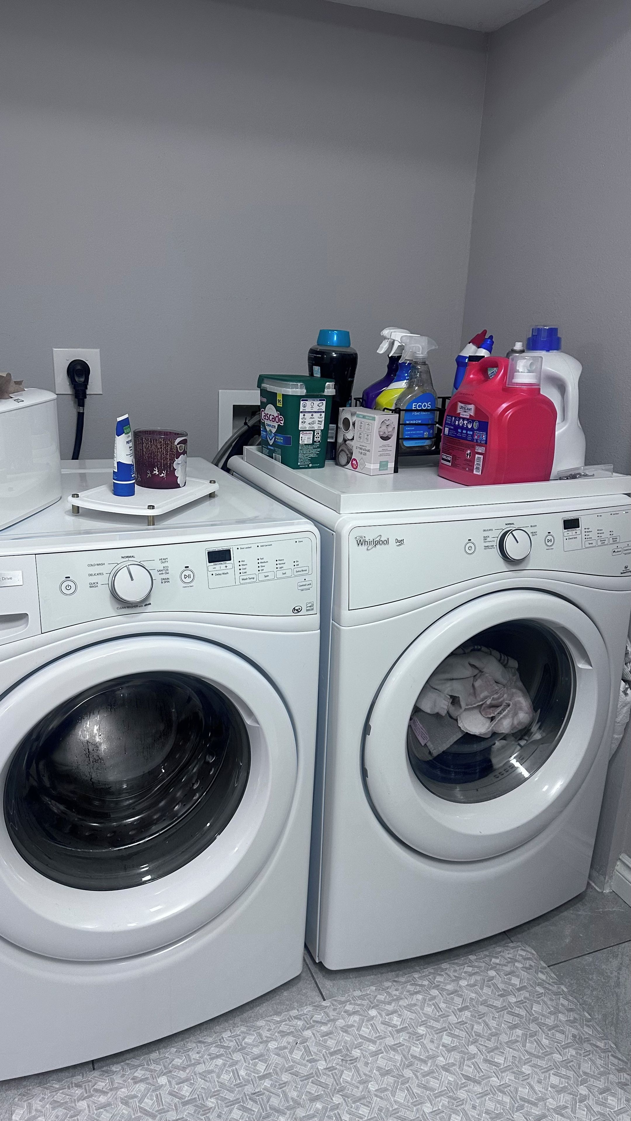 A laundry area with two front-loading Whirlpool washers, a white countertop with various cleaning products, and a grey tiled floor.