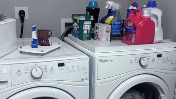 A laundry area with two front-loading Whirlpool washers, a white countertop with various cleaning products, and a grey tiled floor.
