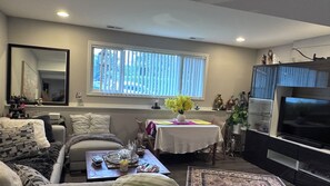 Ein Wohnzimmer mit einer Couch, einem Couchtisch mit Dekoration, einem Esstisch mit Blumen und einem Fernseher, sowie einem Fenster mit Jalousien.