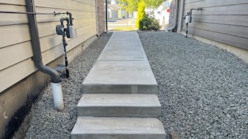 En støbt sti, der fører til et hus med en stenfundament og et metalsystem til nedløbsrør.