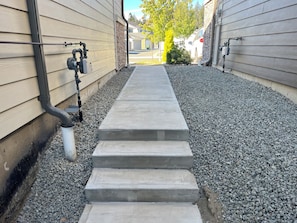 A concrete pathway leading to a house with a stone foundation and a metal gutter system.
