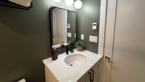 Une salle de bain avec un lavabo blanc, un miroir et un mur vert. Il y a trois lampes au-dessus du miroir et un thermostat sur le mur.