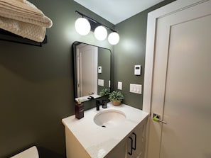 Un baño con un lavamanos blanco, un espejo y una pared verde. Hay tres luces sobre el espejo y un termostato en la pared.