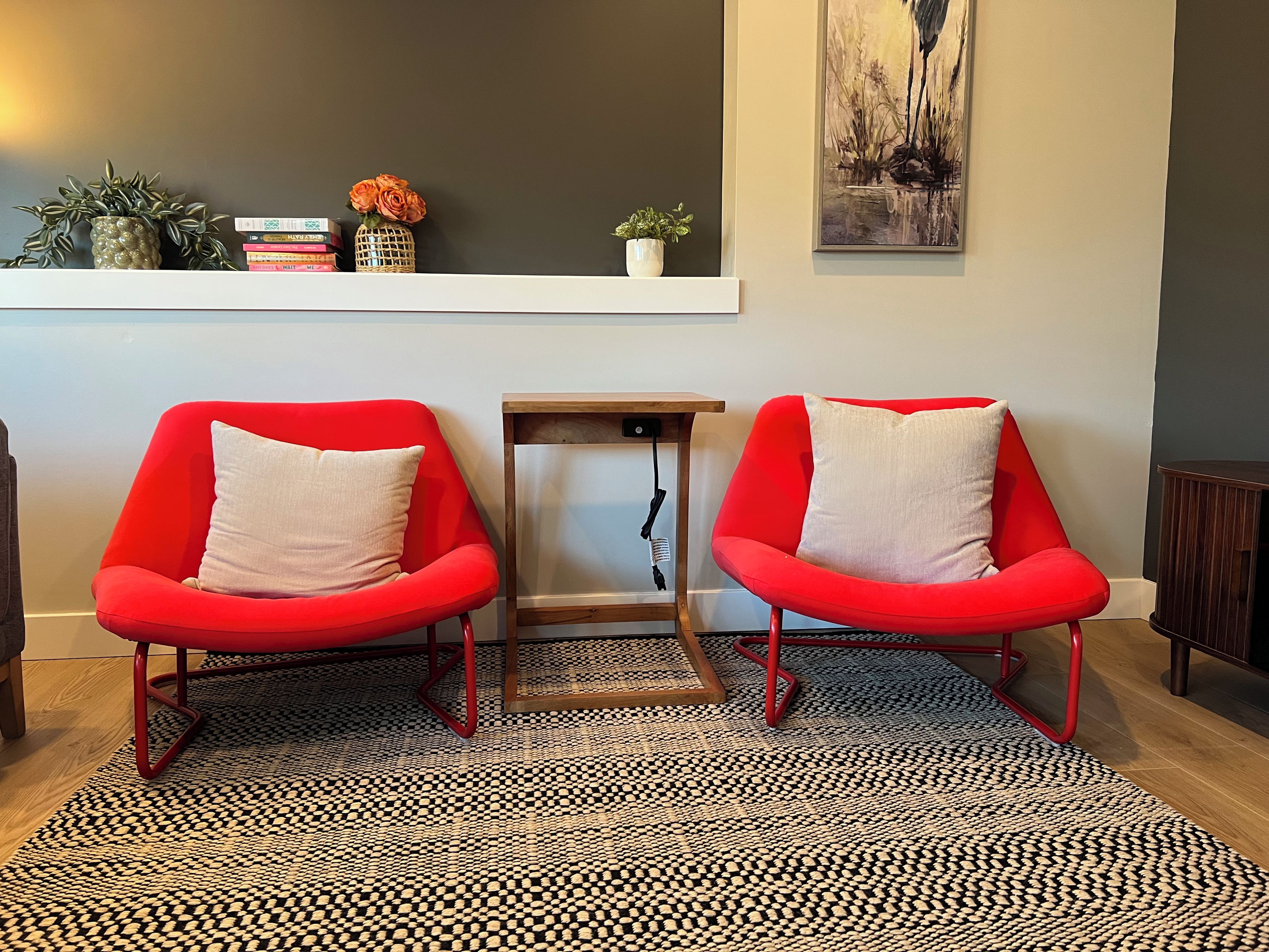 Una habitación con dos sillas rojas, una mesita de madera, un tapete estampado y una estantería con adornos.