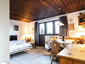 Ein gemütliches Zimmer mit einem Bett, einem Esstisch und einem Stuhl. Ein Fenster mit Vorhängen, eine Uhr an der Wand und eine Lampe sind ebenfalls vorhanden.