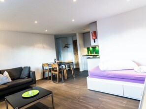 Un salon moderne comprenant un canapé, une table à manger avec des chaises, un petit coin cuisine avec un plan de travail vert, et un lit avec un matelas violet.