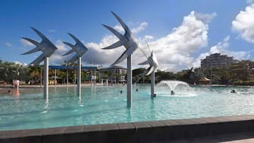 Een zwembad met waterstralen en structuren die doen denken aan windmolens, omgeven door een heldere lucht en een gebouw op de achtergrond.