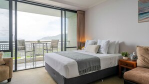 Habitación de hotel con cama, mesita de noche, silla, una pequeña mesa y balcón con vistas.