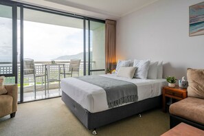A hotel room with a bed, a nightstand, a chair, a small table, and a balcony with a view. - Rydges 92B | 9th Floor City & Esplanade Views (Cairns North)