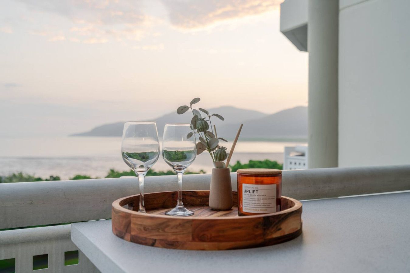 A wooden tray with two glasses of white wine, a small vase with greenery, and a candle labeled 'UPLIFT' on a balcony overlooking a body of water.
