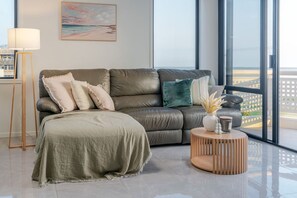 Un salon moderne avec un canapé en cuir, une petite table d’appoint ronde en bois et une vue sur la plage grâce à de grandes fenêtres.