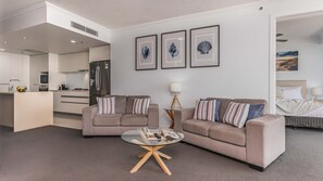 Ein modernes Wohnzimmer mit einer Couch, einem Couchtisch und einem Bett im Hintergrund. An den Wänden hängen gerahmte Bilder, und im Hintergrund ist eine Küchenzeile erkennbar.