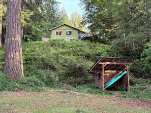 Una casa rodeada de un bosque denso, una estructura de madera con un tobogán y un área cubierta de césped.