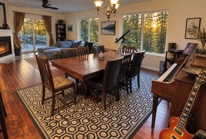A spacious living room with a dining area, a fireplace, a piano, and large windows with a forest view. - Private Mission Lake retreat: hot tub, dock, paddleboards & record player (Bremerton)