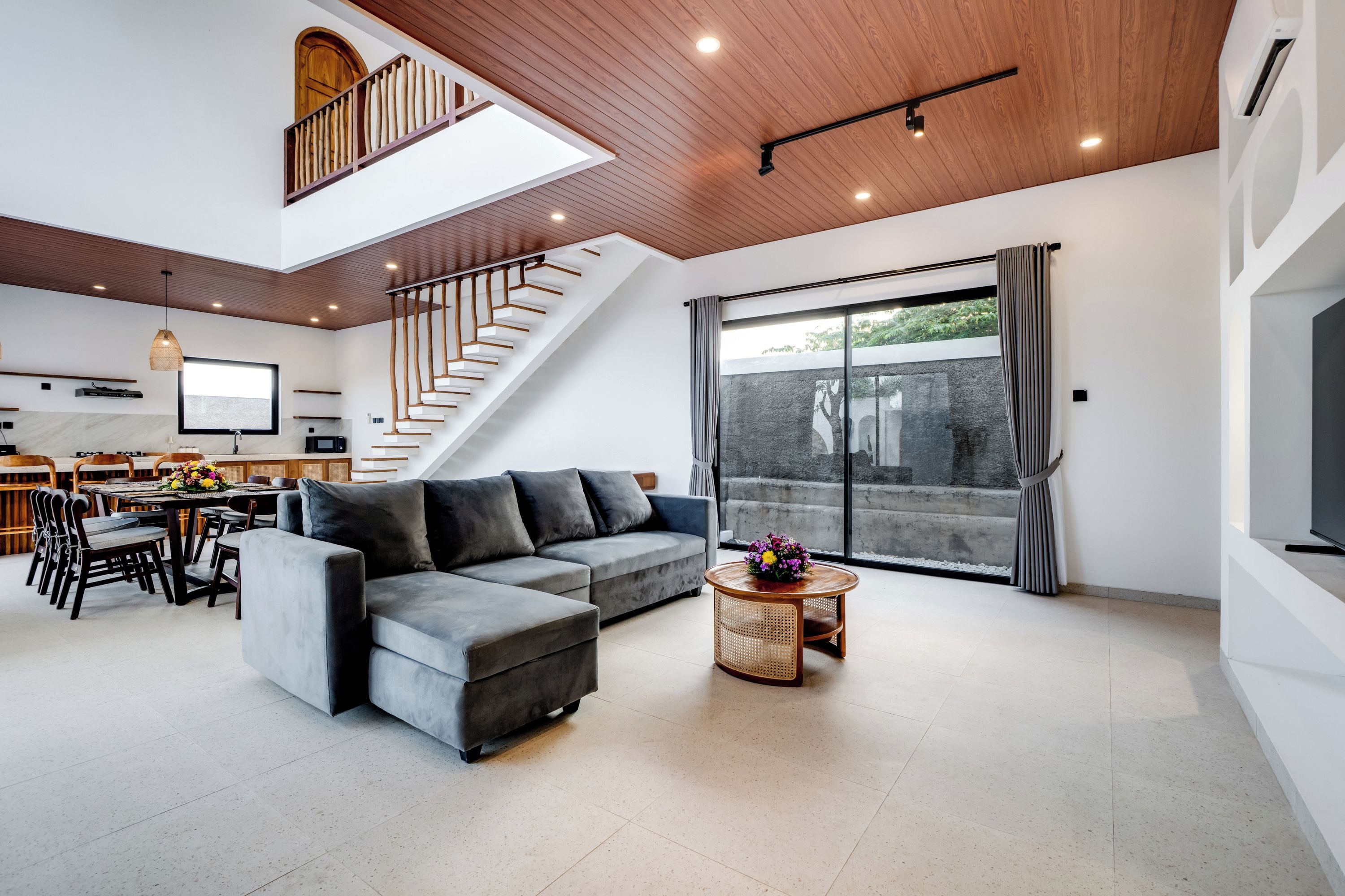 A spacious living area with a modern staircase, a grey sectional sofa, a dining table with chairs, and a large sliding glass door.