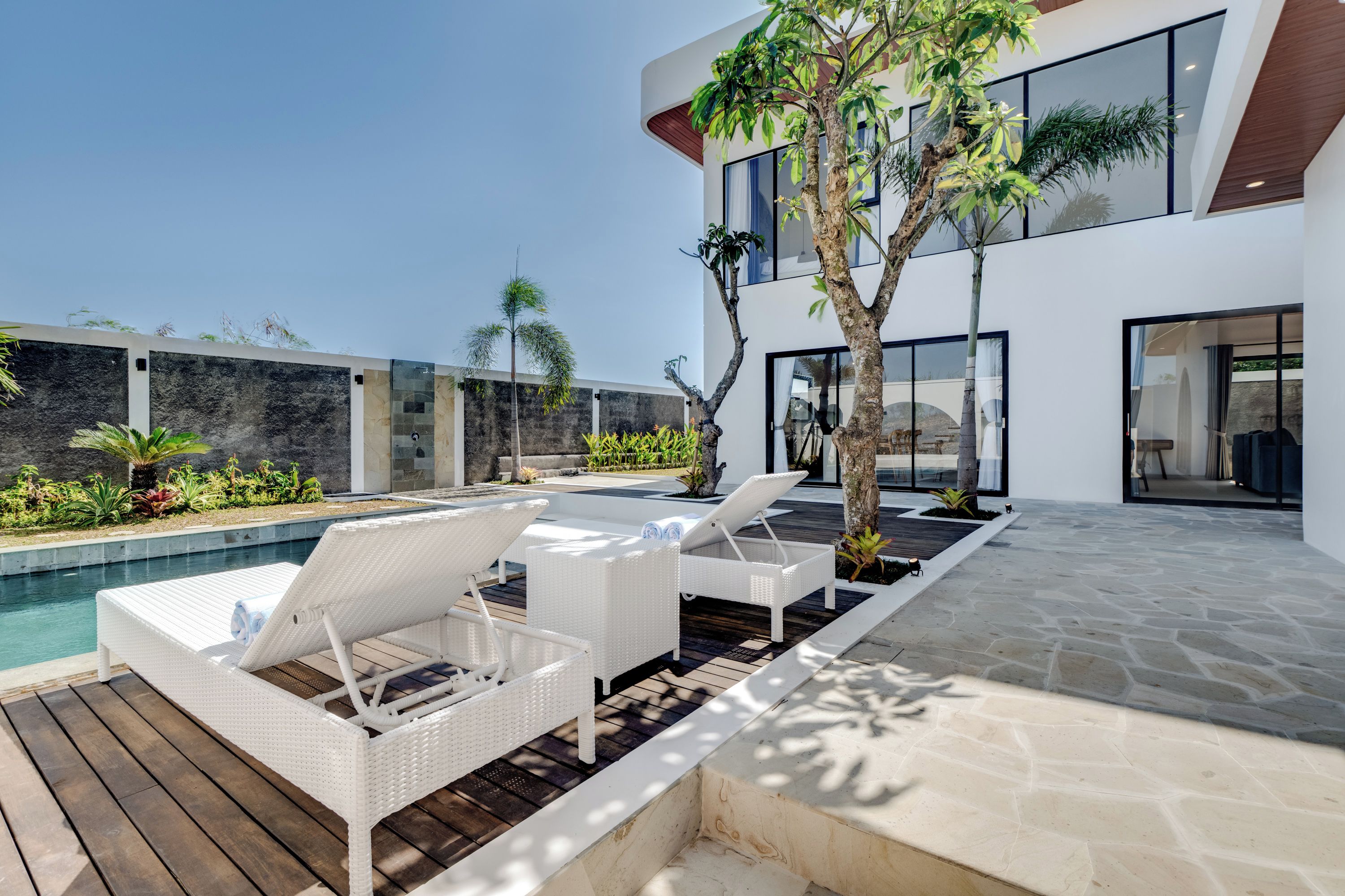 Un área de piscina moderna al aire libre con sillones de mimbre blancos y una terraza de piedra.