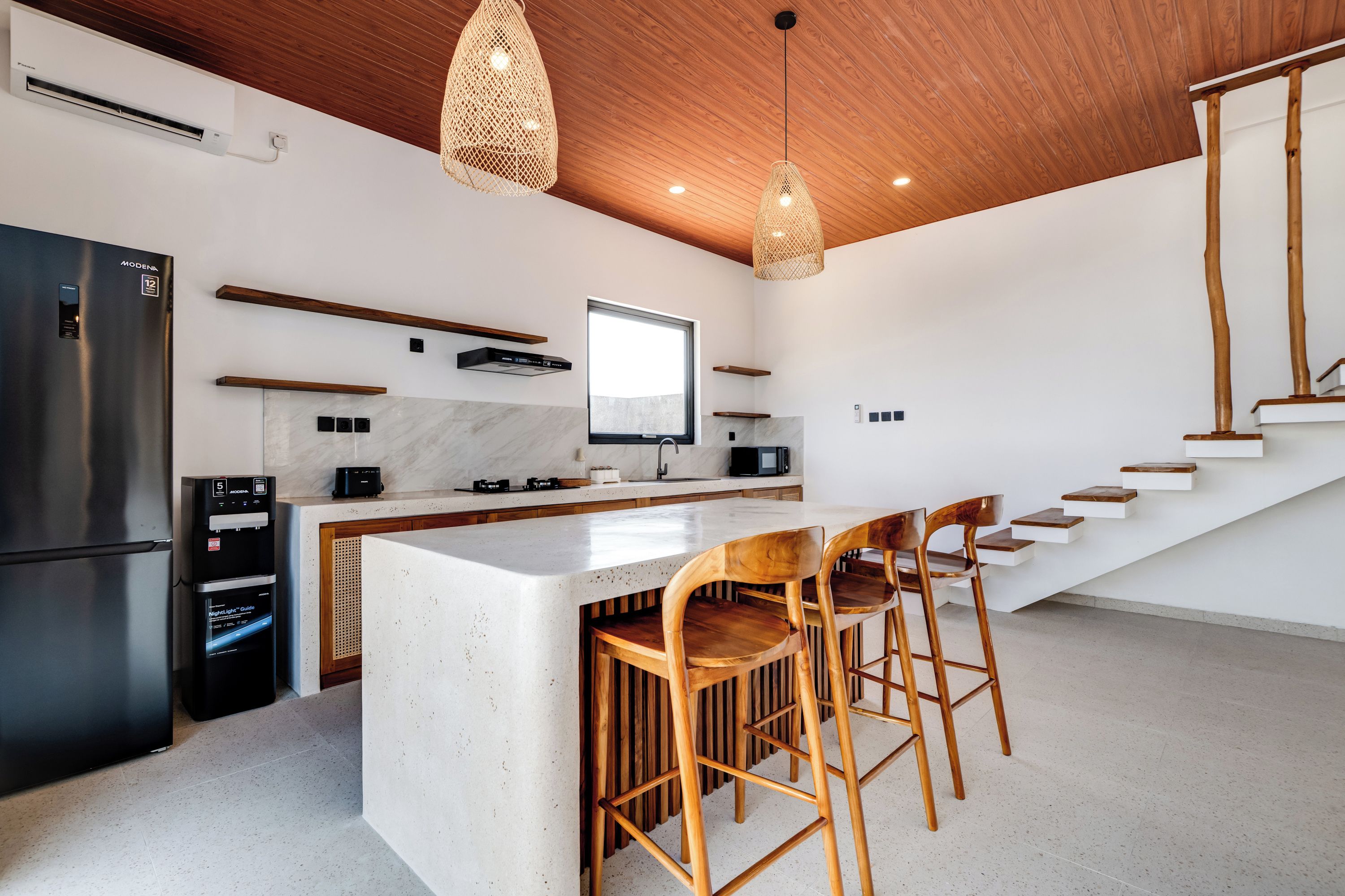 Una cocina moderna con una isla central, taburetes de madera, refrigerador empotrado y una escalera que sube a un nivel superior.