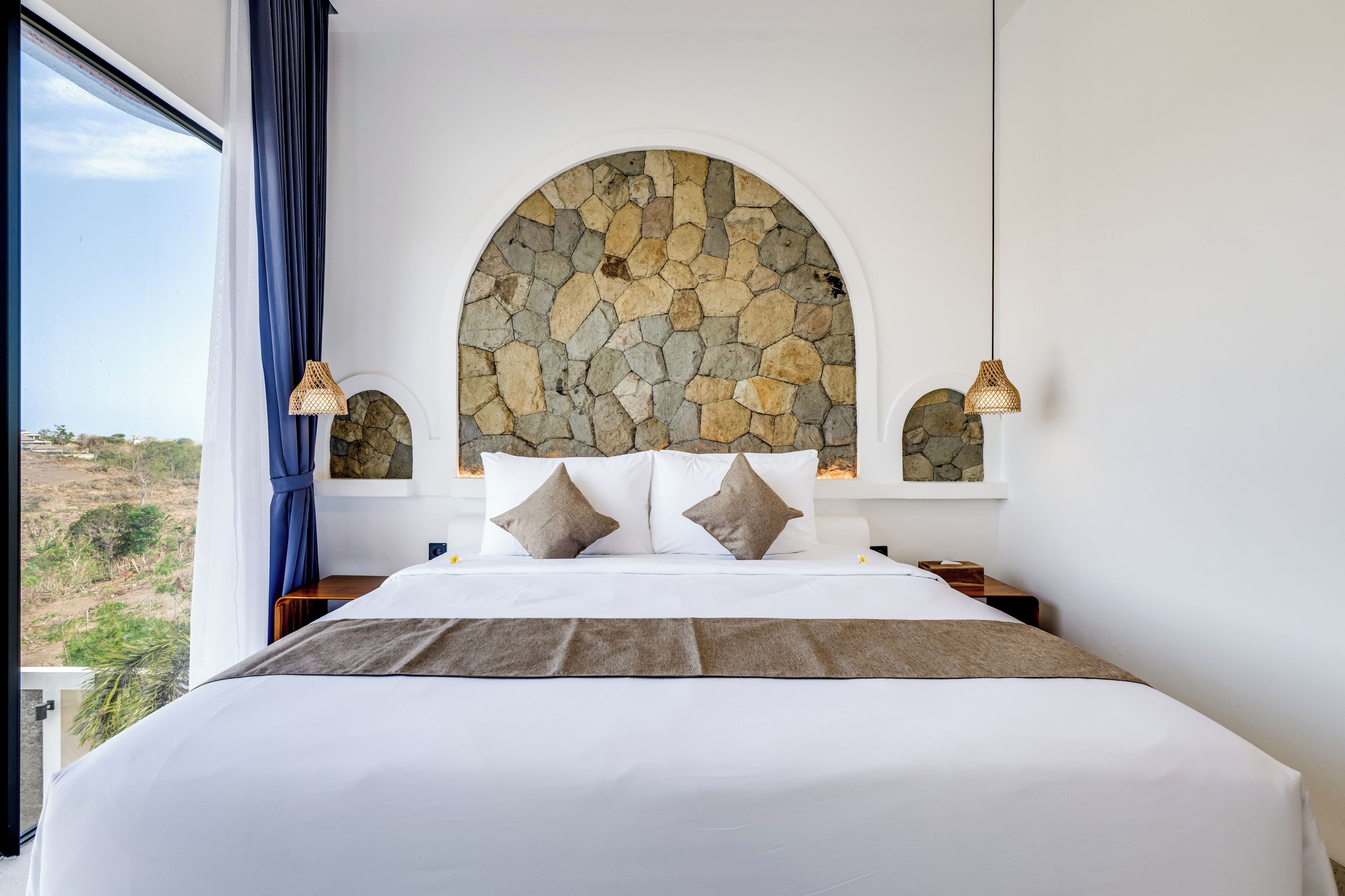Ein Schlafzimmer mit einer Steinkonstruktion, einem großen Bett mit weißer Bettwäsche, zwei Nachttischlampen und Blick ins Freie.