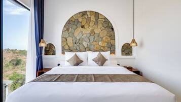 Ein Schlafzimmer mit einer Steinkonstruktion, einem großen Bett mit weißer Bettwäsche, zwei Nachttischlampen und Blick ins Freie.
