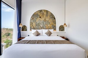 A bedroom with a stone wall arch, a large bed with white bedding, two bedside lamps, and a view of the outdoors. - Samanea Hills (Bali)
