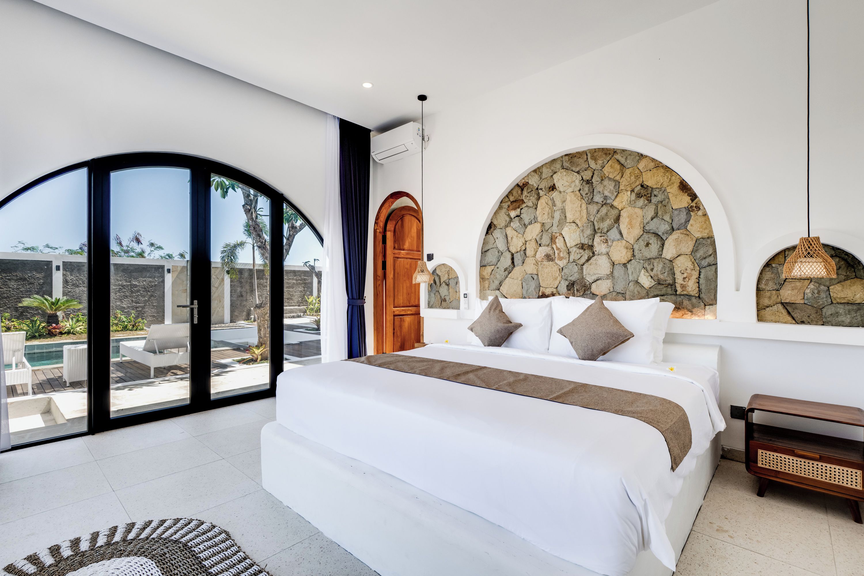 Un dormitorio moderno con una cama grande, una pared de piedra y vista a una piscina y zona exterior.