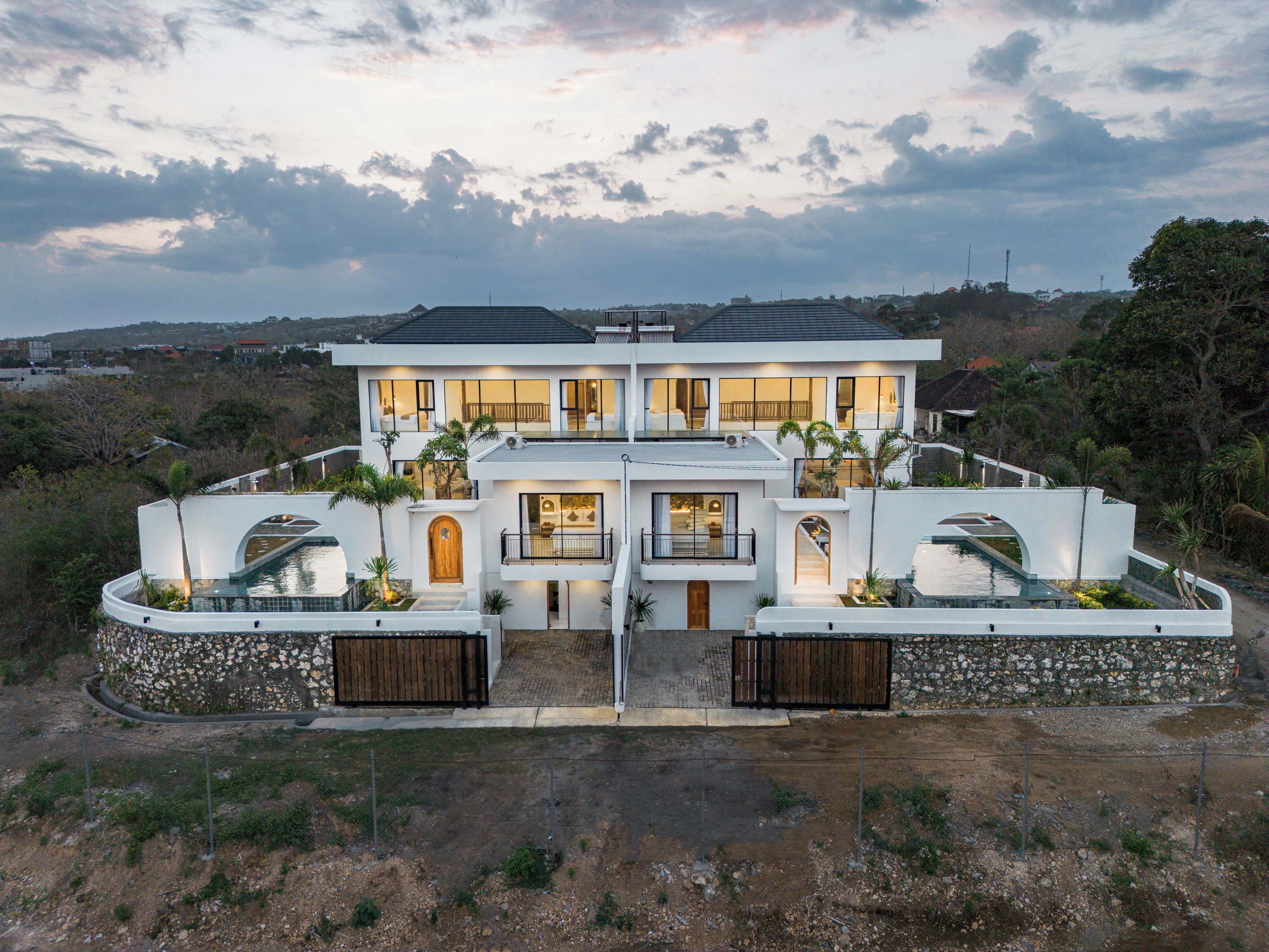 Una casa moderna de dos pisos con piscina, rodeada por un muro de piedra y palmeras.