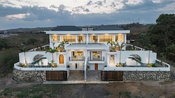 Una casa moderna de dos pisos con piscina, rodeada por un muro de piedra y palmeras.