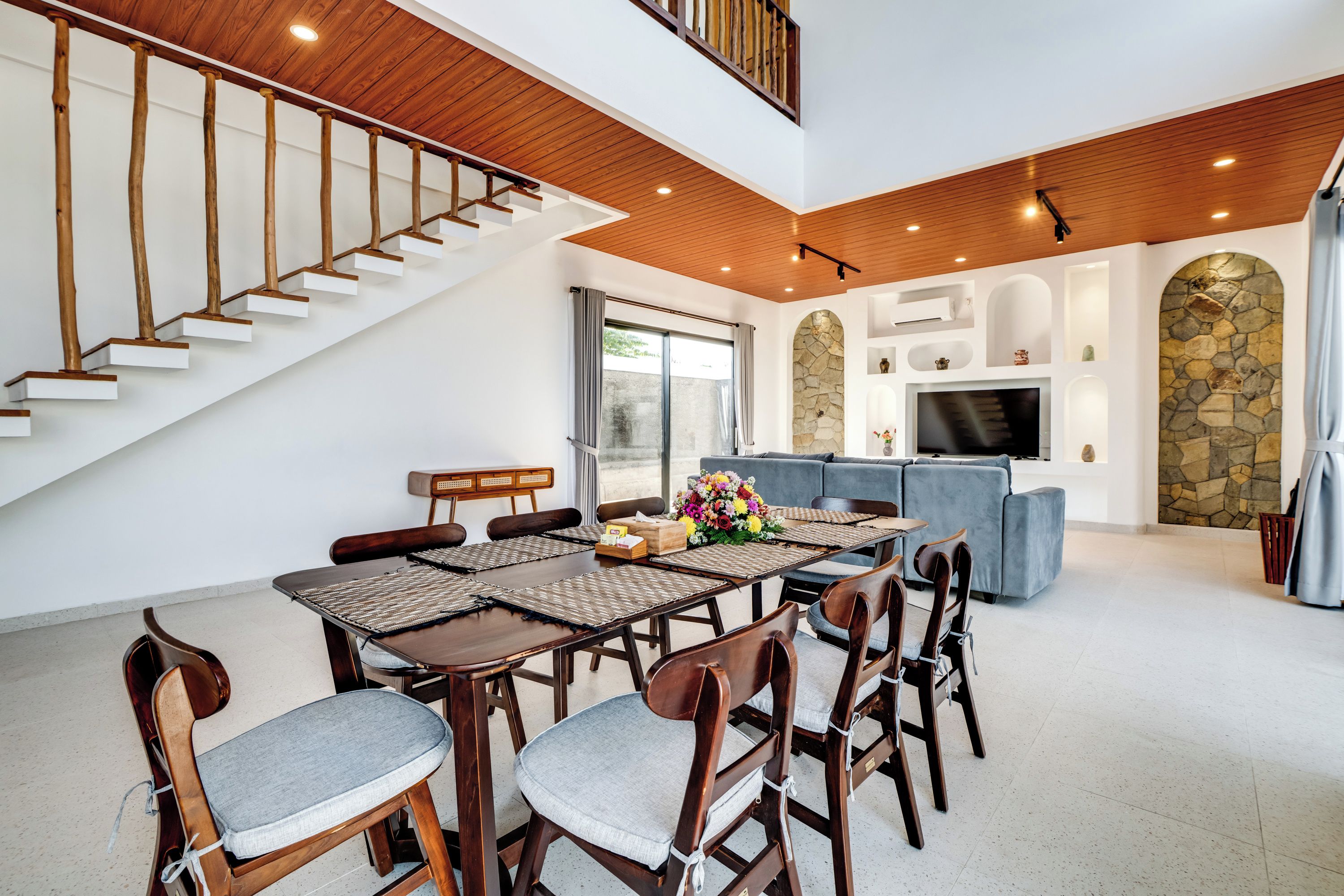 A spacious living area with a dining table set for six, a sofa, and a television mounted on a stone wall.