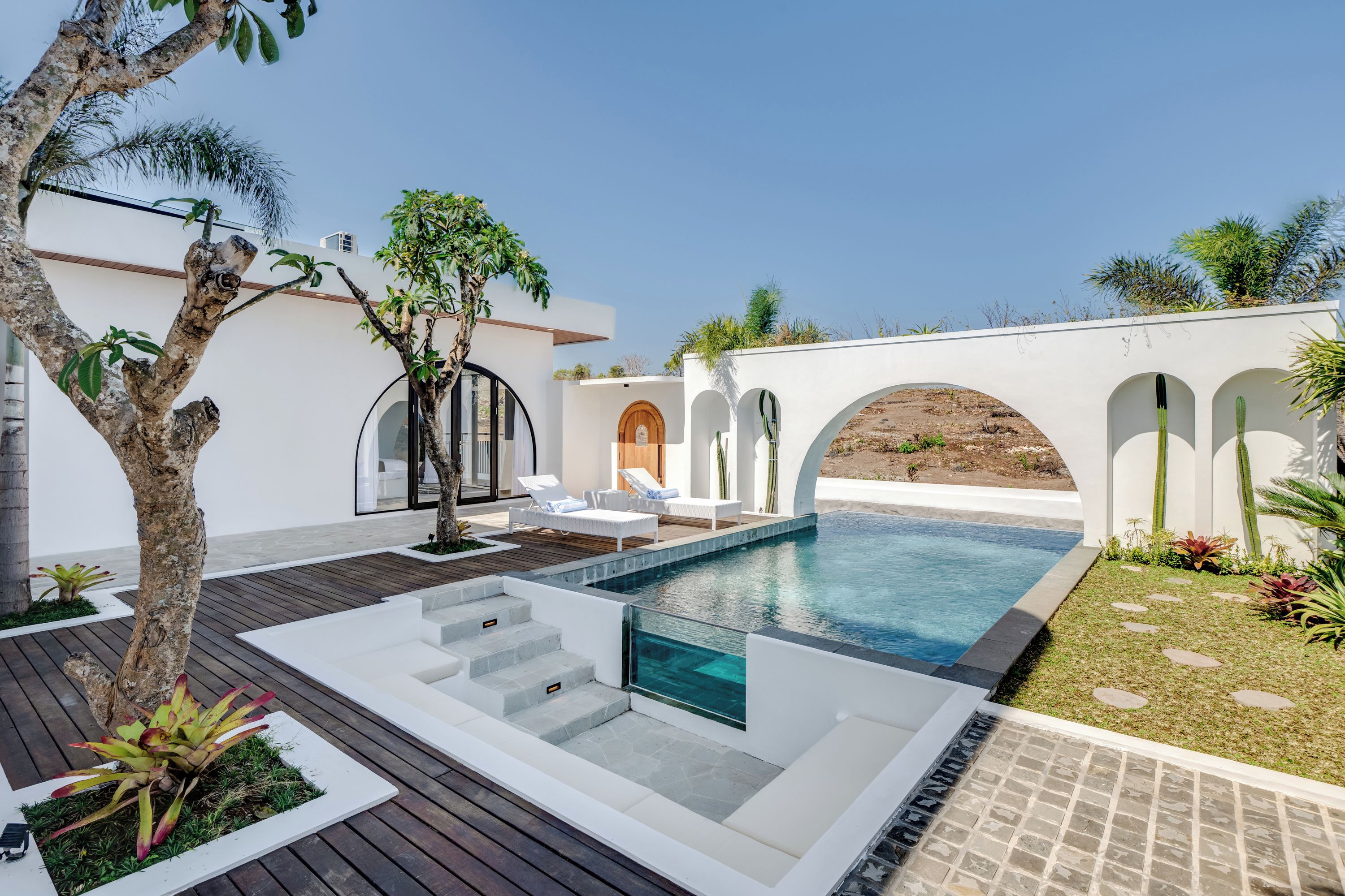 Un área de piscina exterior moderna con un deck de madera, sillas de descanso y una pared blanca con arcos.