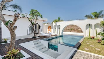 Un área de piscina exterior moderna con un deck de madera, sillas de descanso y una pared blanca con arcos.