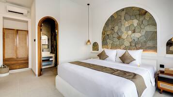 Un dormitorio con una cama grande, un arco de pared de piedra, un armario de madera y un baño visible a través de un arco.