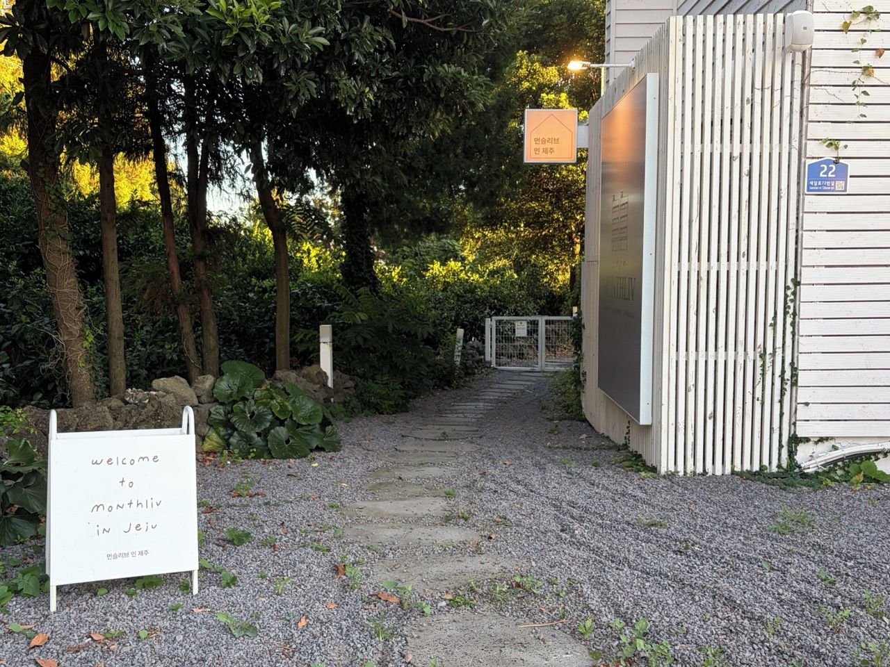 Uma trilha de cascalho leva a um edifício com uma placa que diz 'Welcome to Monthliv in Jeju'.