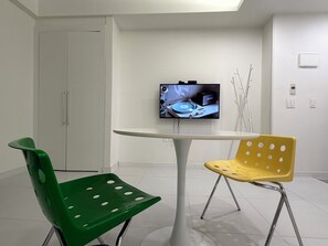 Una habitación moderna con una mesa blanca, dos sillas, un televisor en la pared y una puerta.