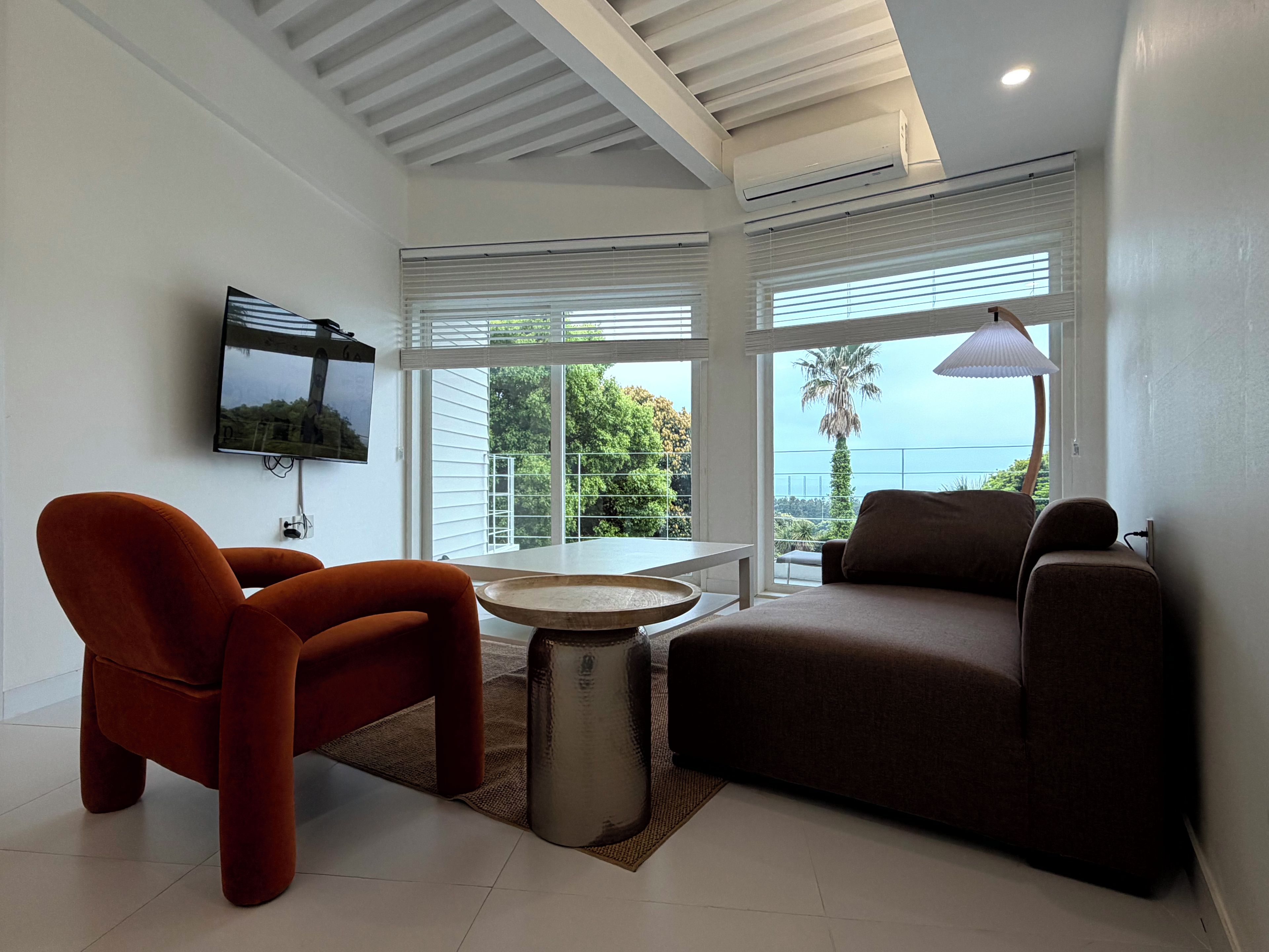 A modern living room with a flat-screen TV, two sofas, a coffee table, and large windows with blinds.