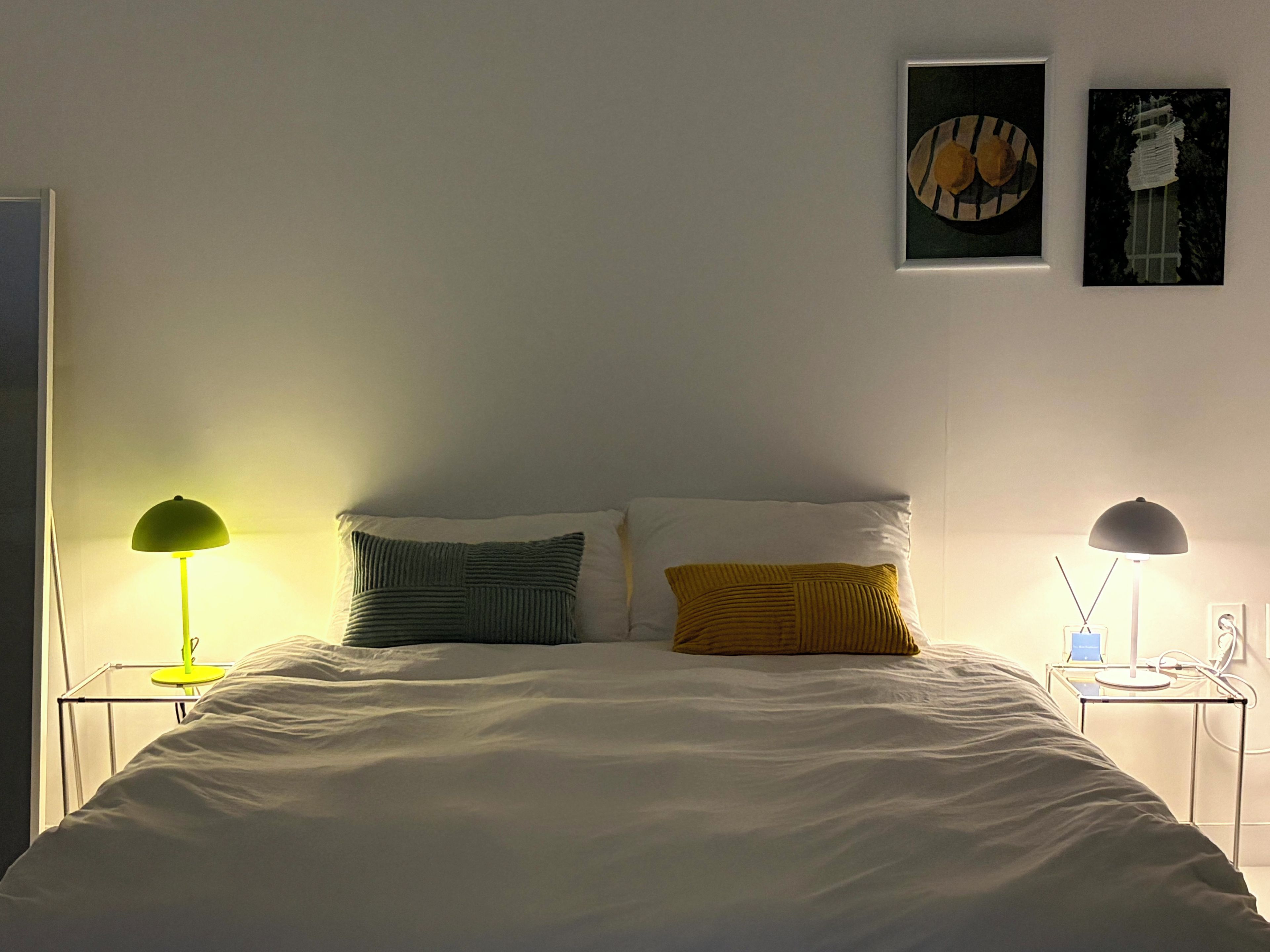 A neatly made bed with two pillows, a bedside table with a lamp, and two wall-mounted artworks.