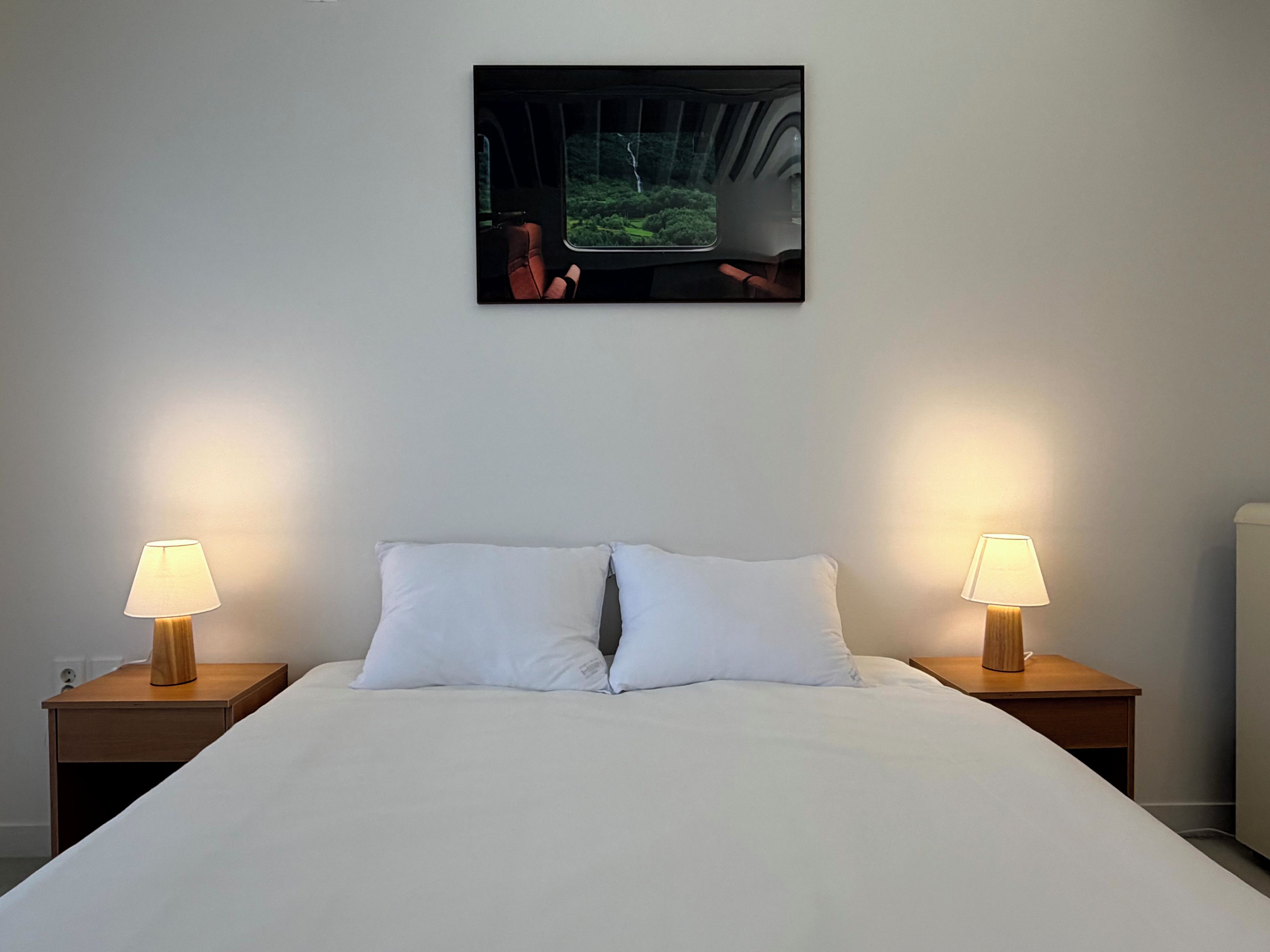 Una habitación de hotel con una cama, dos lámparas de noche, un cuadro en la pared y una mesita de noche.