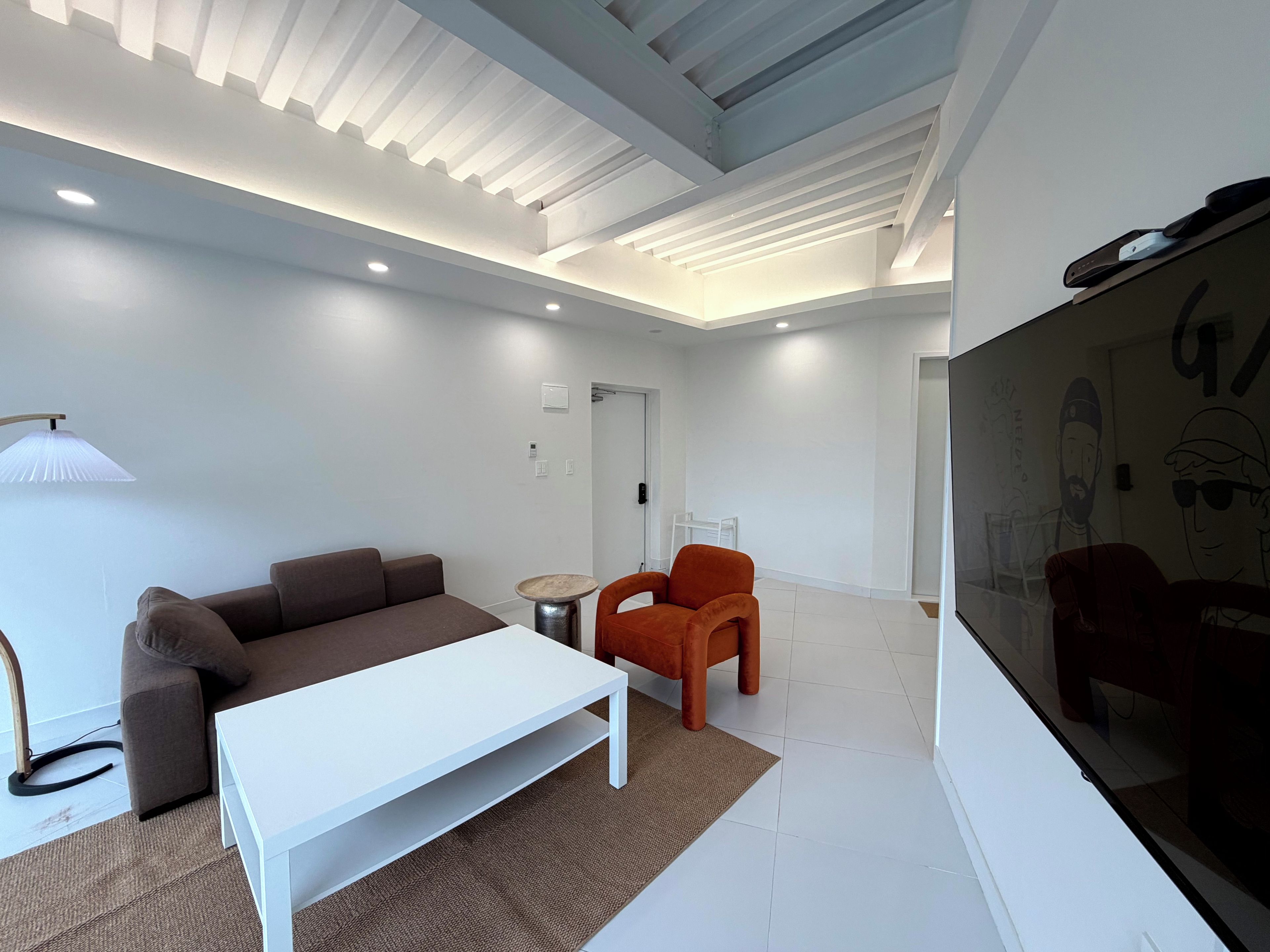 A modern living room with a white sofa, a white coffee table, and an orange chair. There is a lamp on the left, a small table with a round top, and a large TV screen on the right.
